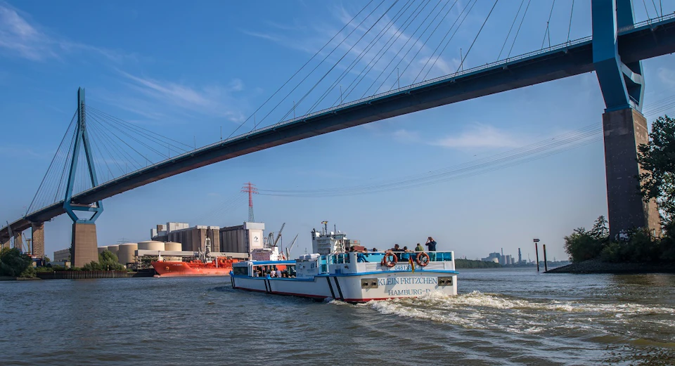 A ship labeled "Kleiner Fritzchen Hamburg" on a river, with an industrial area and bridge in the background under a clear blue sky.