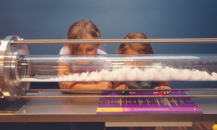 Dvě děti pozorují pěnové vzory uvnitř průhledného válce na stolním displeji.