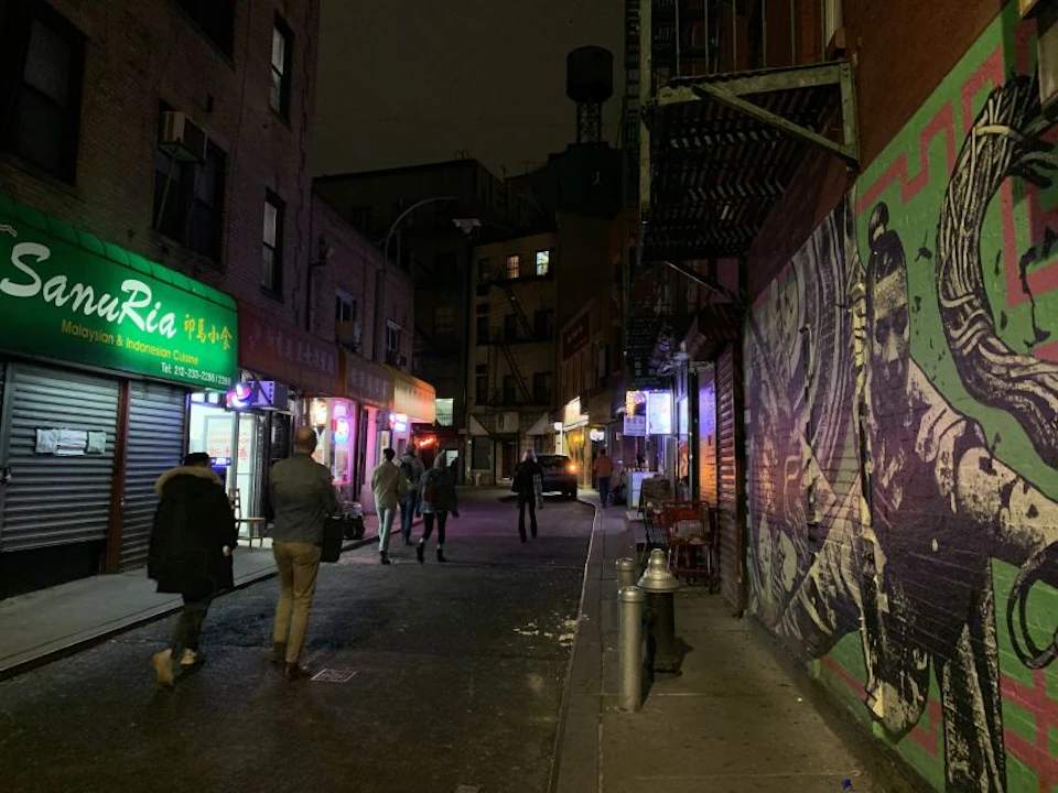 People walking in a narrow alley at night, with neon-lit storefronts and graffiti murals on the walls.