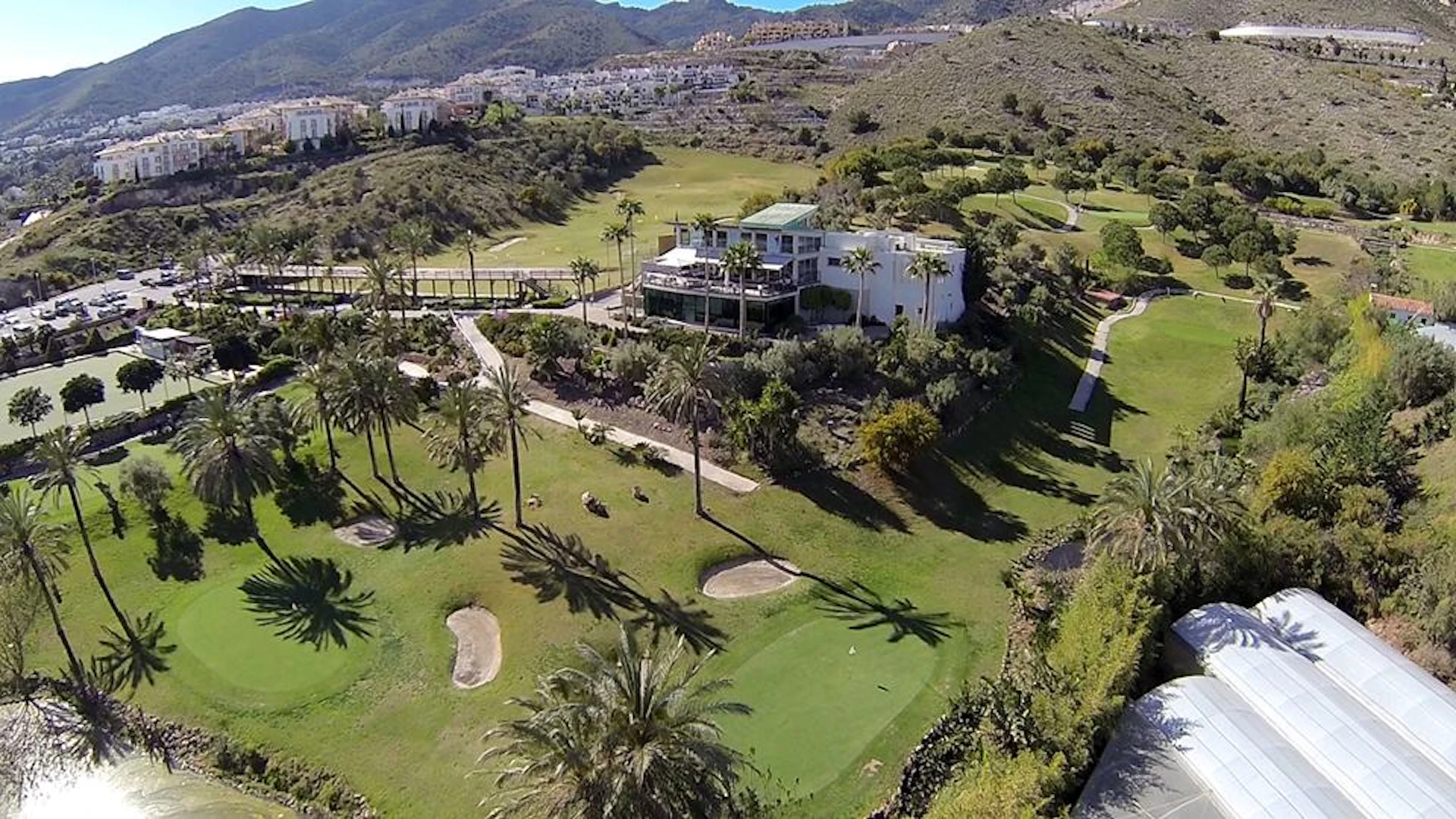 Aerial view of a golf course with a modern building surrounded by trees and hills, with distant buildings and mountains in the background.