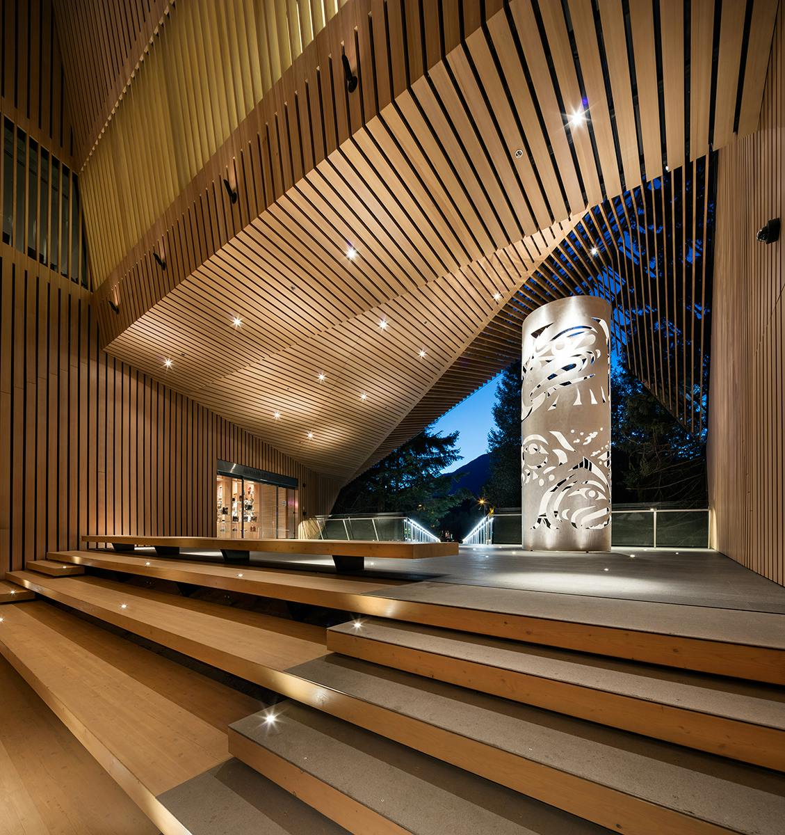 Espace intérieur moderne avec des panneaux en bois, des escaliers éclairés, un balcon, de grandes fenêtres et une colonne décorative.