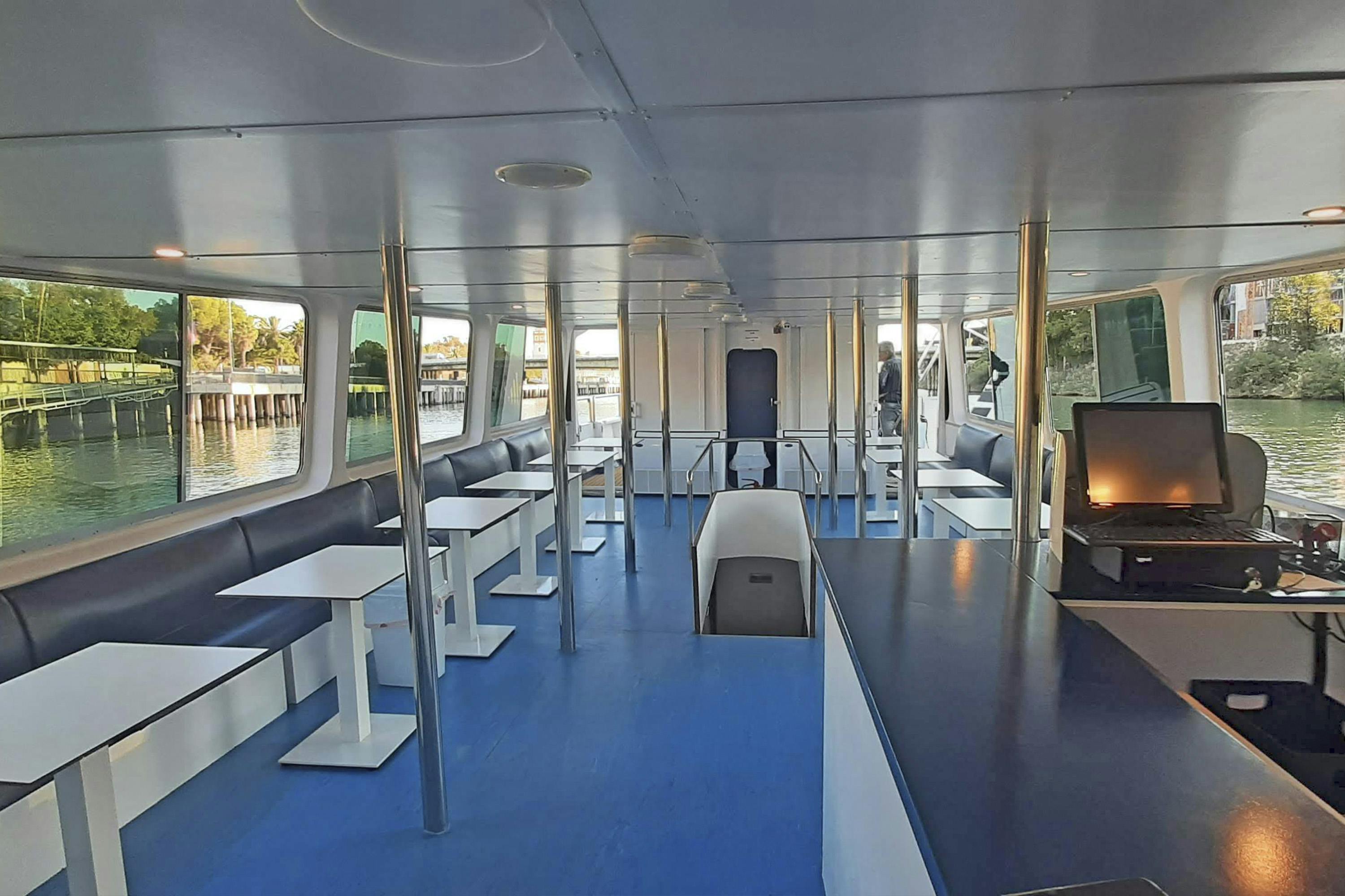 Wnętrze przestronnej, nowoczesnej łodzi z niebieską podłogą, białymi stołami i niebieskimi ławkami, z dużymi oknami i metalowymi słupkami.