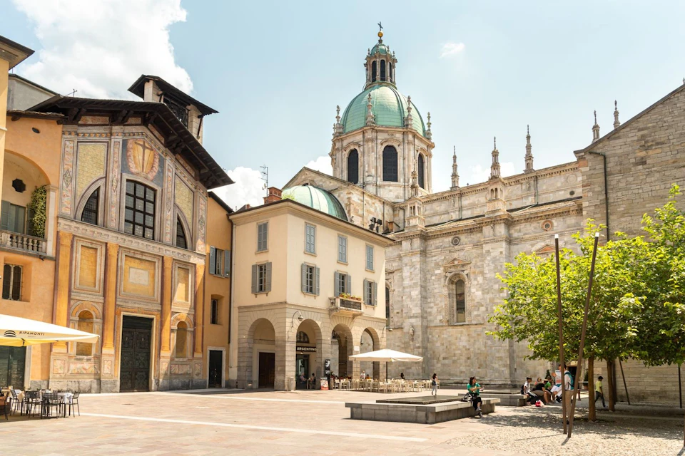 A historic square with a cathedral, colorful buildings, a tree, and people sitting and walking under a sunny sky.