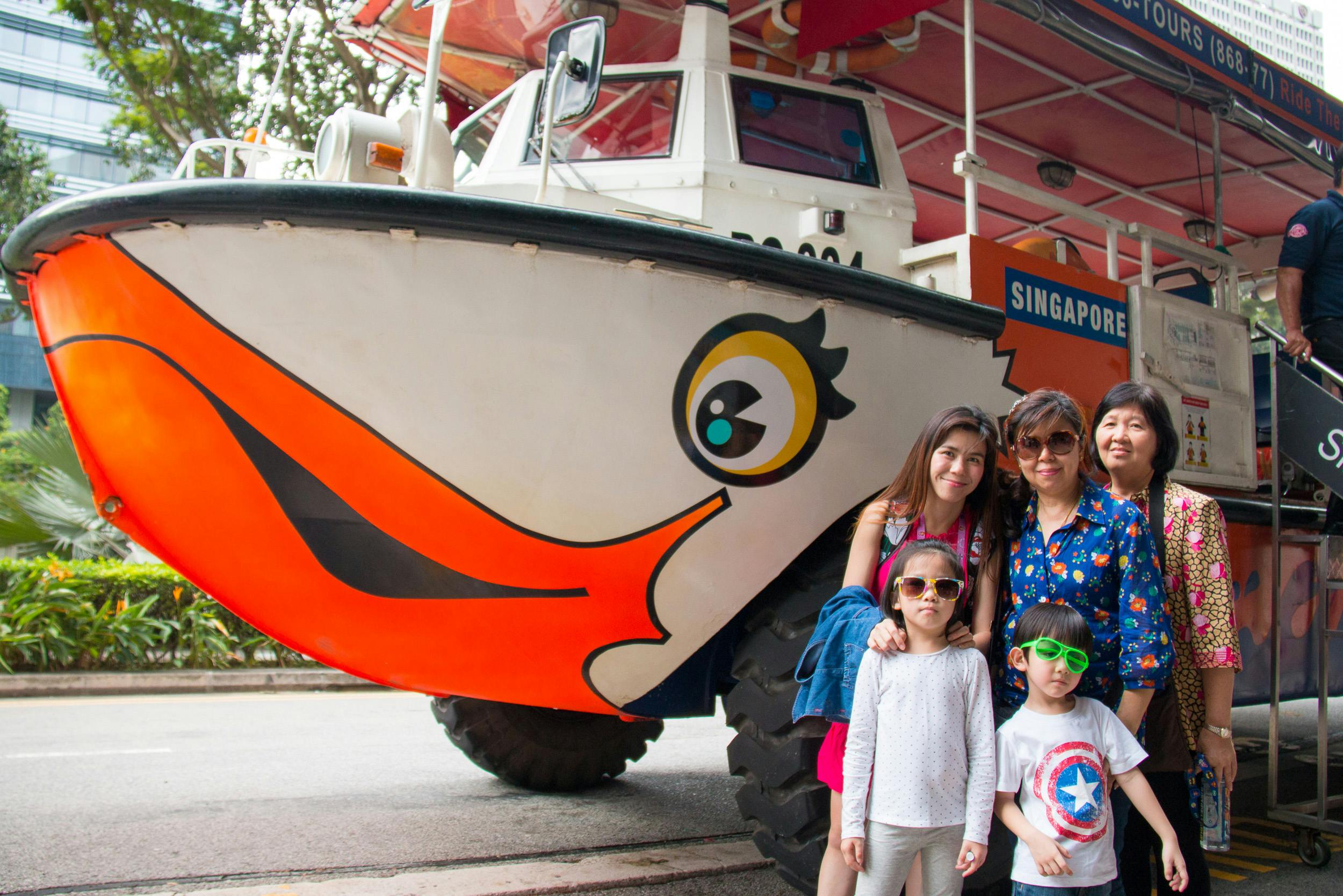 Четыре человека позируют перед лодкой с нарисованным оранжевым клювом и глазом. На борту лодки написано «Сингапур».