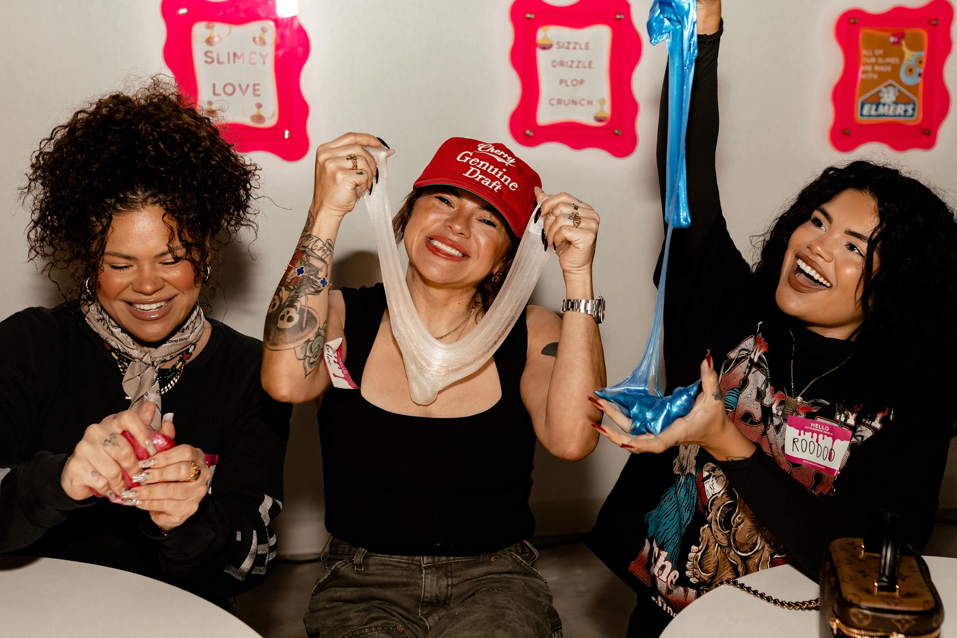 Two women smiling and playing with slime; one wears a red cap, the other wears a scarf. Bright signs with text in the background.