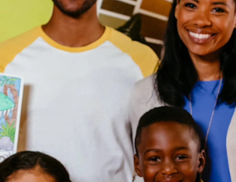 A smiling family of four holding craft items. They are in front of a colorful wall with creative elements.