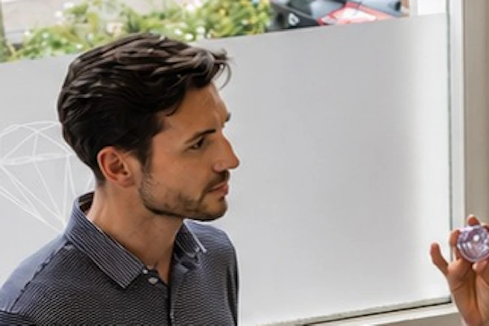A group of people standing indoors near a window, with one person explaining something while holding a small object.