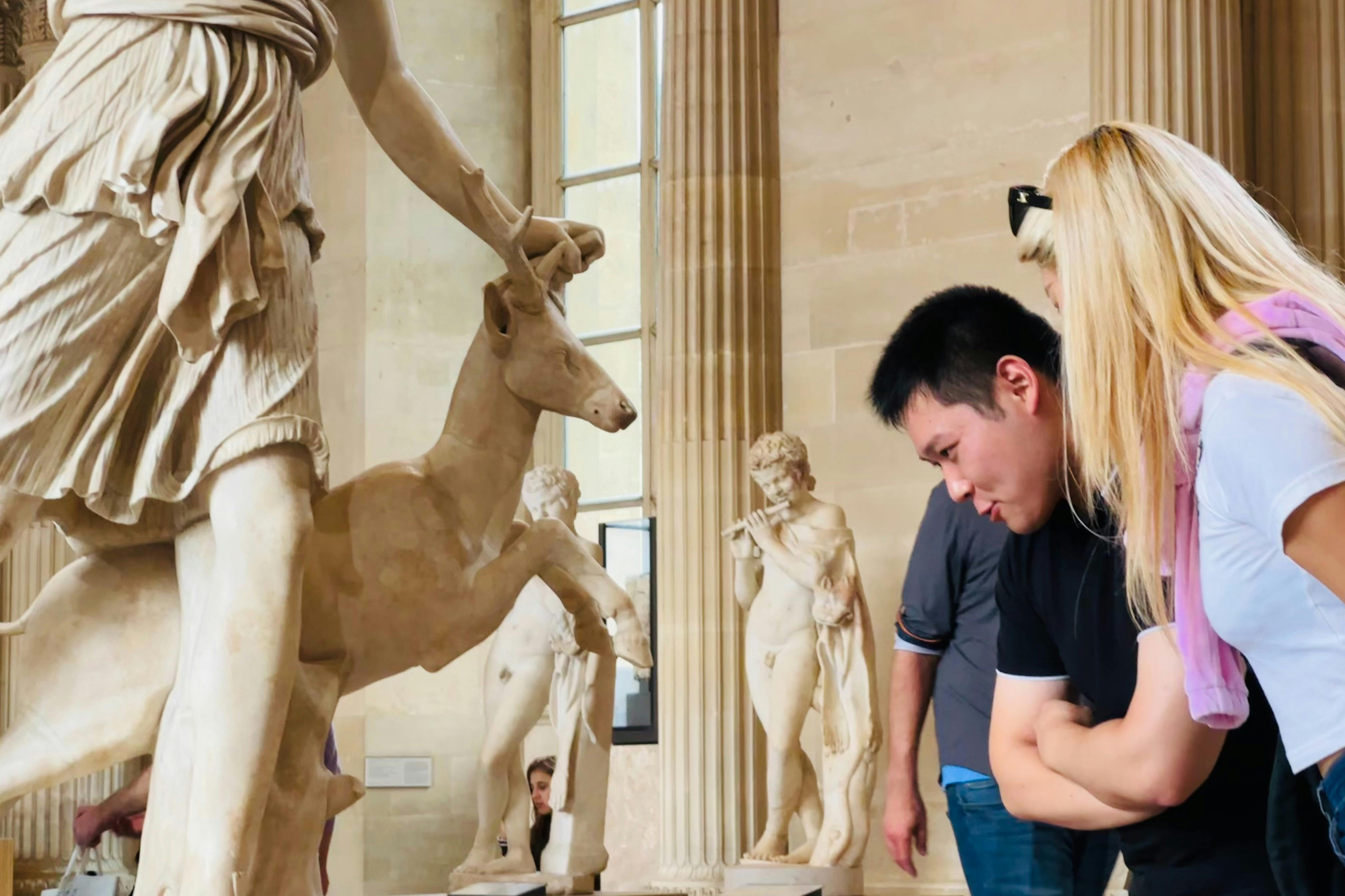 두 사람이 박물관에서 사슴과 플루트를 연주하는 사람의 모습을 포함한 고전 동상을 면밀히 관찰하고 있습니다.