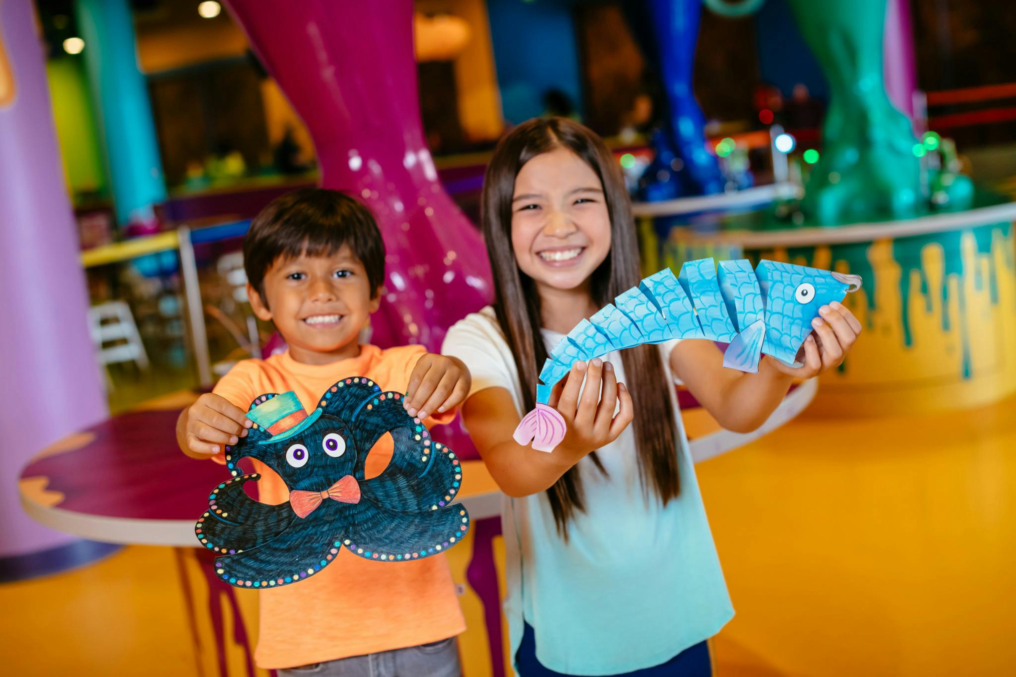 Two smiling children holding colorful paper crafts in a vibrant indoor setting with playful decor and bright lights.