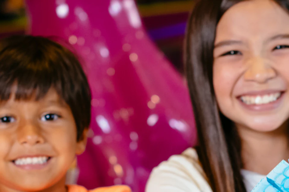 Two smiling children holding colorful paper crafts in a vibrant indoor setting with playful decor and bright lights.