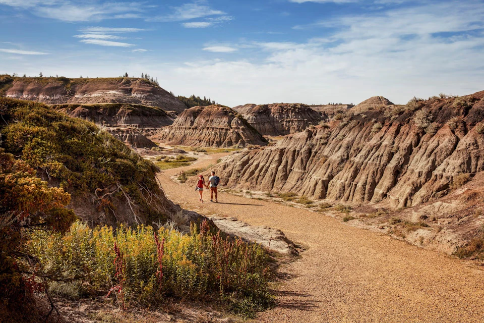 Two people walking on a dirt path amidst rugged, dry rock formations under a blue sky with light clouds.