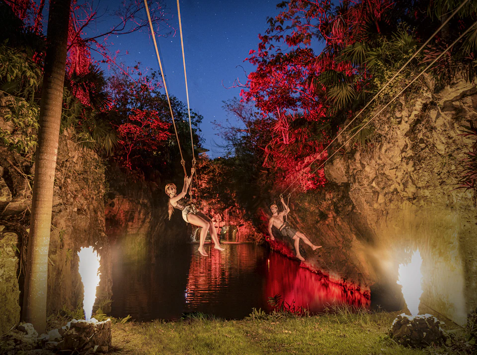 Two people zip-lining over a waterway, surrounded by rocky terrain and trees illuminated with red lights, with fire on each side.