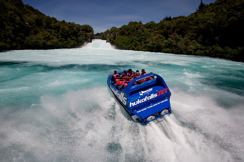 A blue jet boat with passengers speeds through a river toward a waterfall, surrounded by lush greenery under a blue sky.