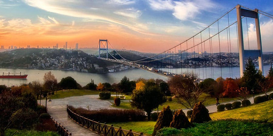 Suspension bridge over a river with heavy traffic, surrounded by city buildings and greenery under a colorful sunset sky.