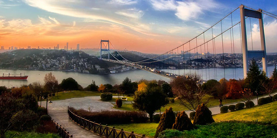 Suspension bridge over a river with heavy traffic, surrounded by city buildings and greenery under a colorful sunset sky.