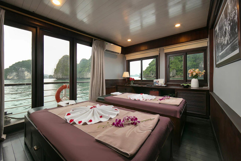 A room with two massage tables adorned with towels and flowers, large windows showcasing a scenic water view with greenery.