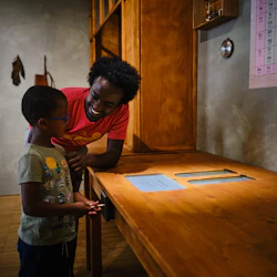 Ett barn i en grön skjorta och en man i en röd skjorta som ler mot varandra i ett rum med ett skrivbord i trä och ett periodiskt bord på väggen.