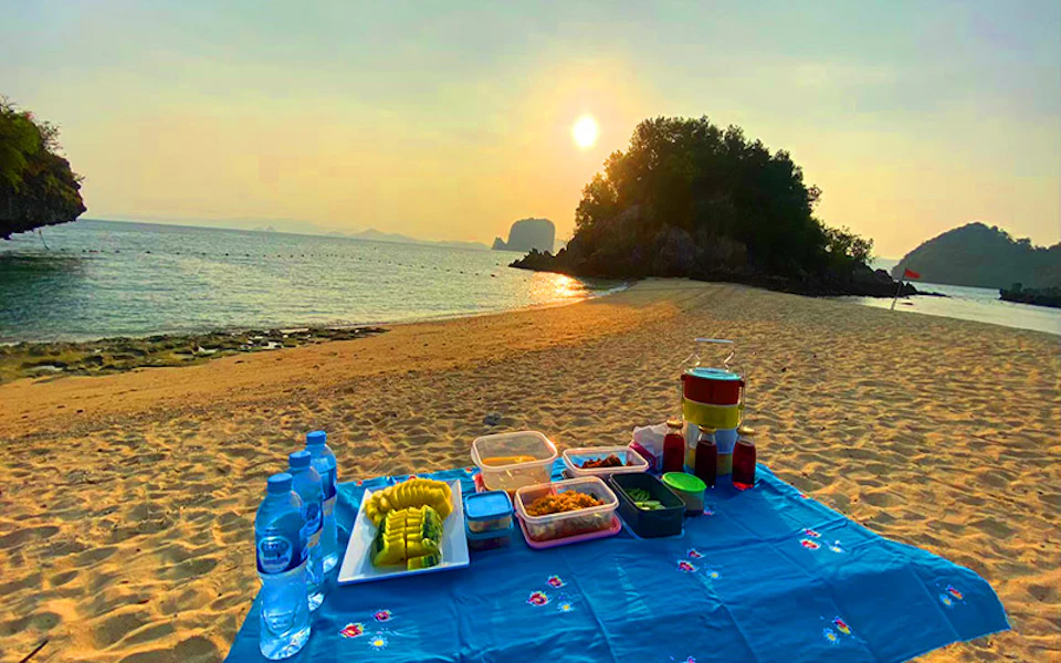 A variety of food and drinks arranged on a blue cloth on a sandy beach at sunset, with water and a small island in the background.