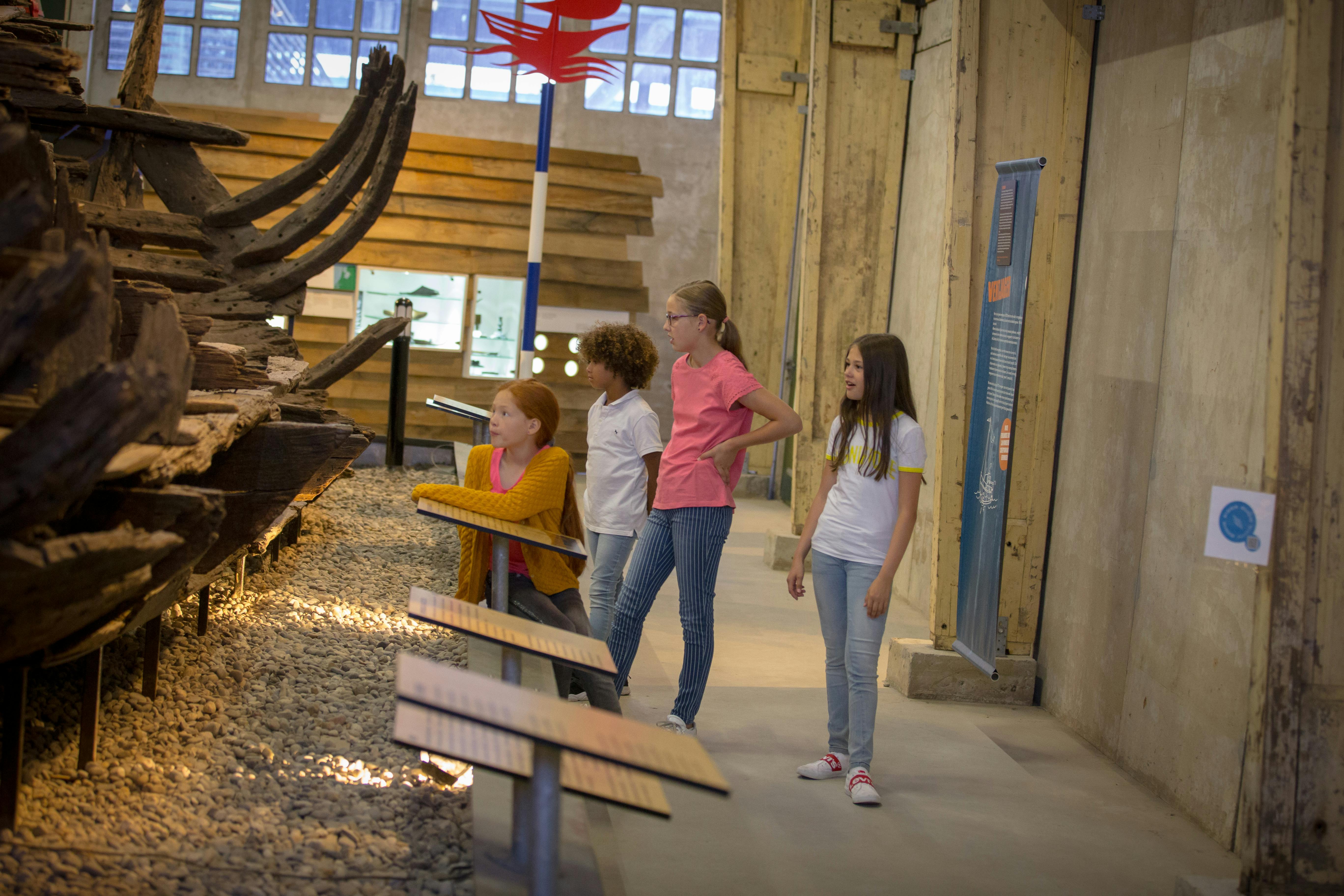 Čtyři děti stojí v muzeu a pozorují starobylou dřevěnou loď obklopenou informačními panely.