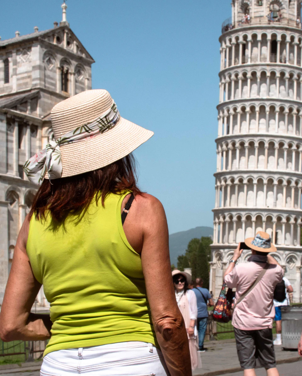 A person in a green top and hat observes the Leaning Tower of Pisa, while others take photos in the background.