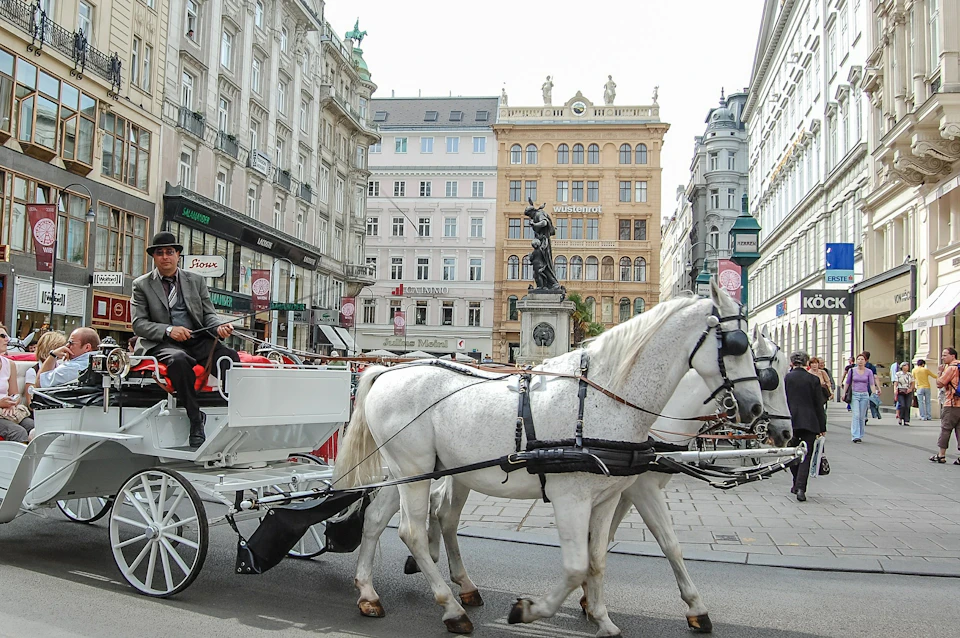 A horse-drawn carriage with a driver and passengers moves through a bustling city street lined with historic buildings.