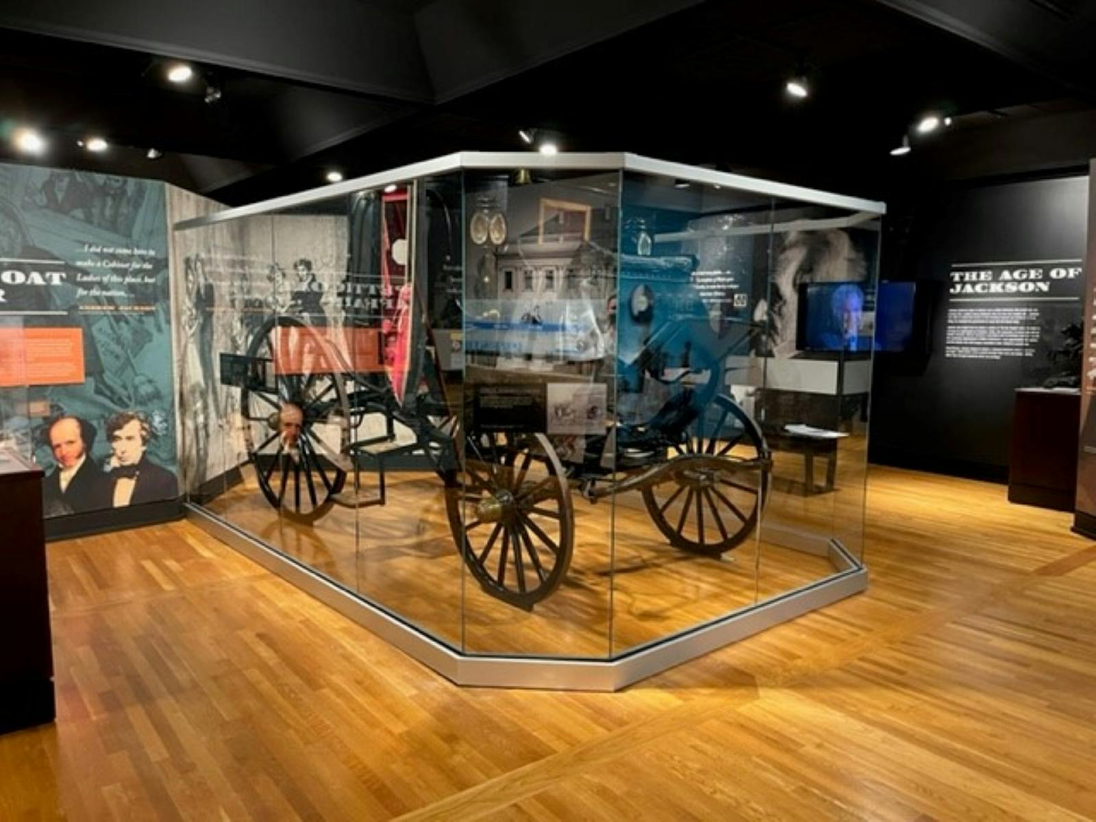 Museum exhibit with a vintage carriage encased in glass, informational displays, wooden floor, and historical photographs on the walls.