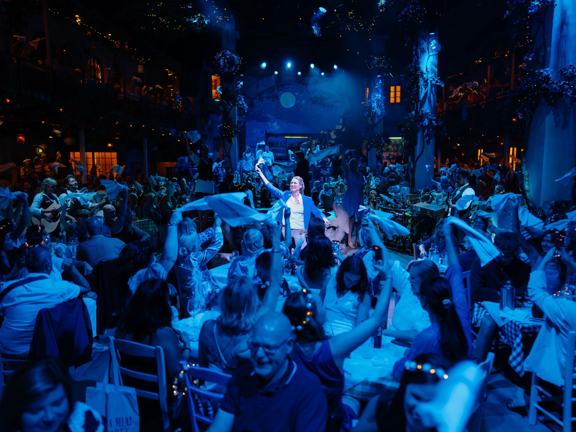 A lively indoor event with a crowd of people waving napkins, colorful lighting, and a performer standing in the center.