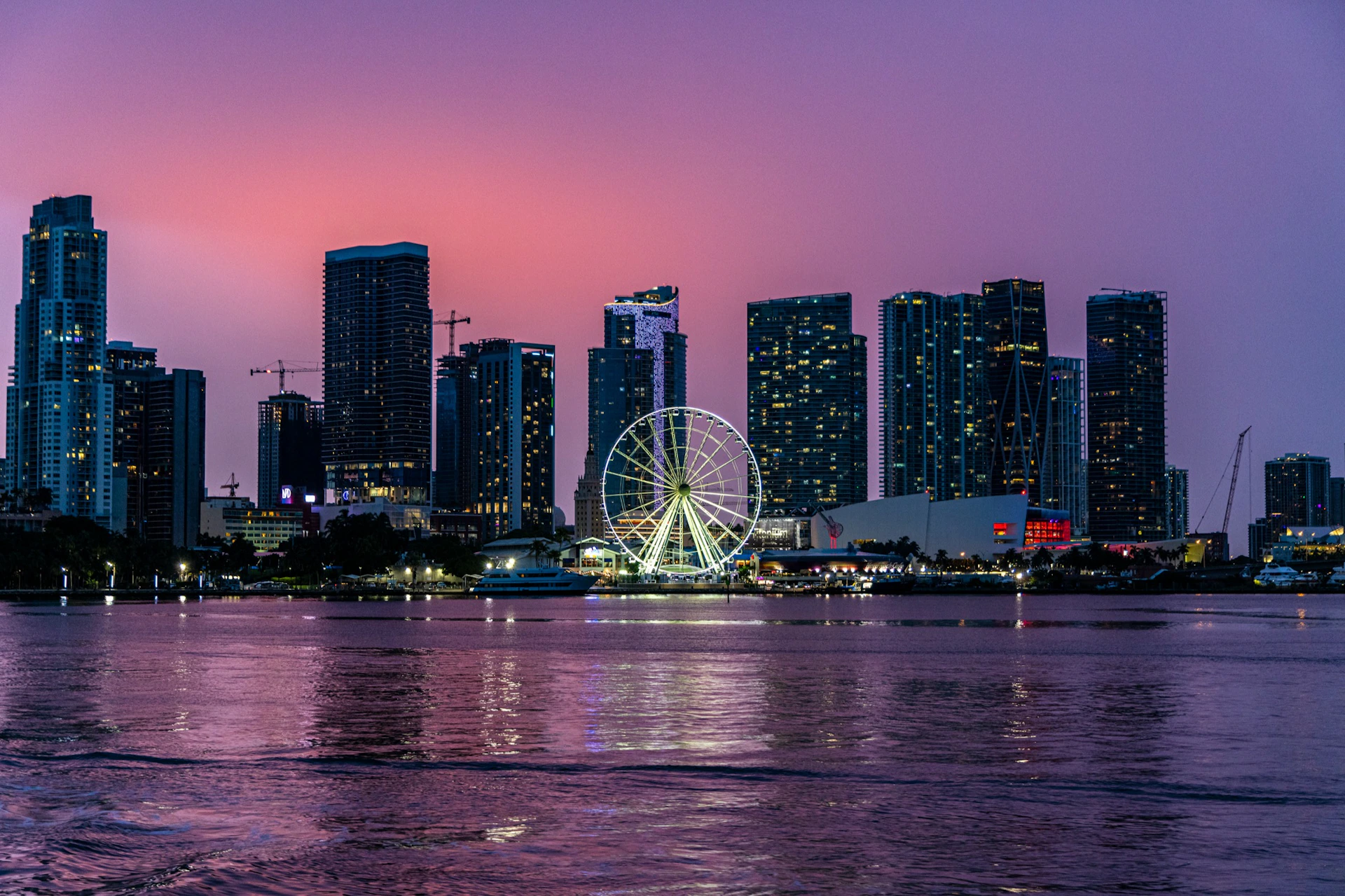 City skyline at sunset with tall buildings, a Ferris wheel, and reflections on the water in the foreground.