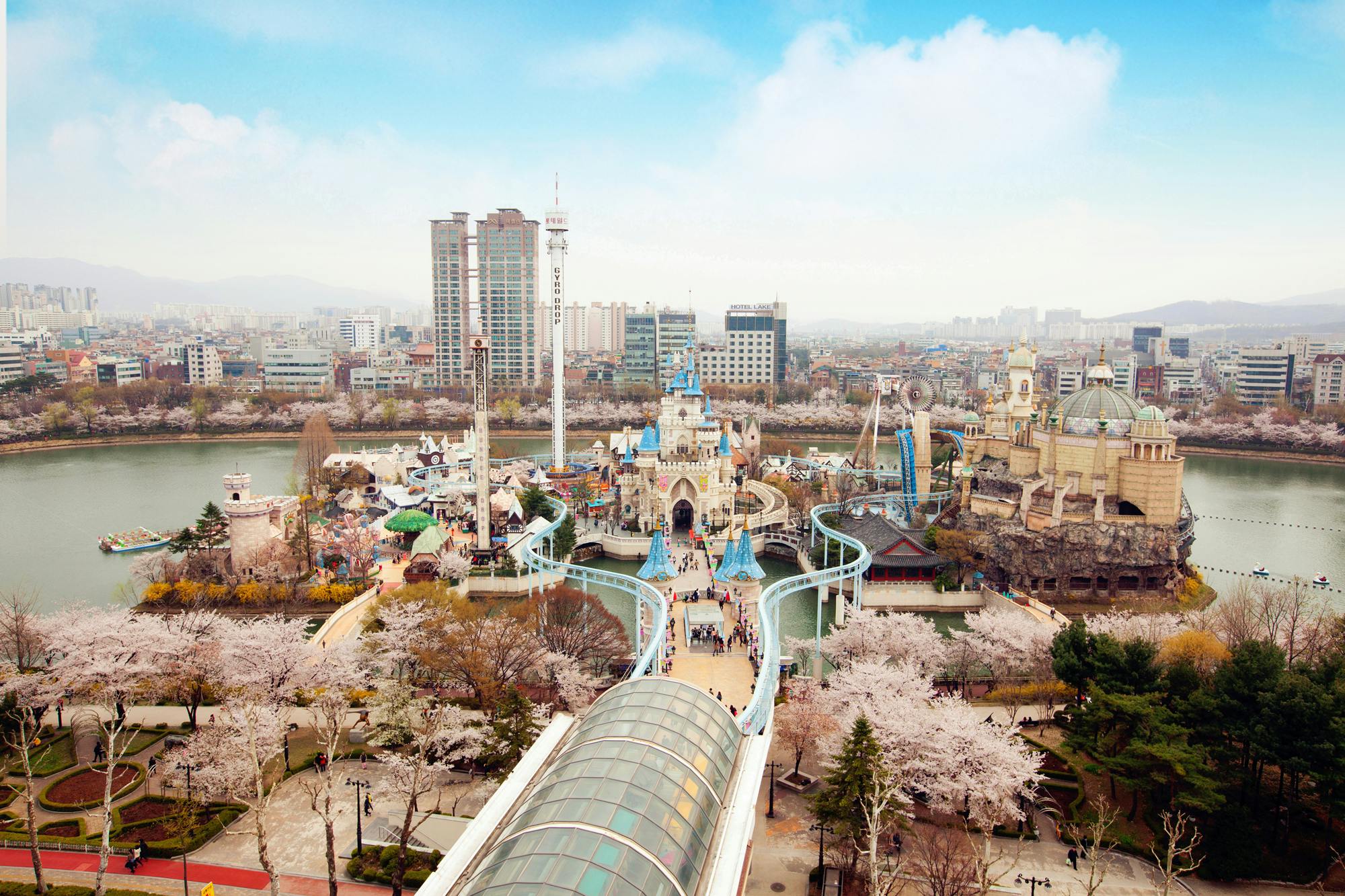 Aerial view of a theme park with water rides, castle structures, and surrounding cherry blossom trees, set against a backdrop of city buildings.