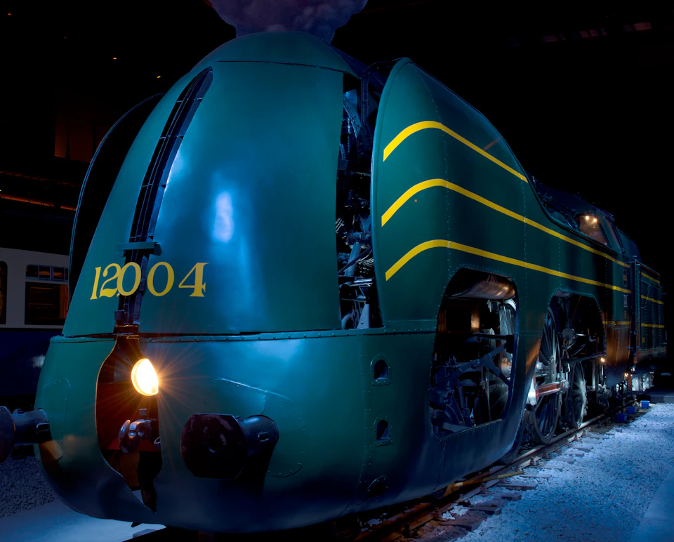 A green vintage steam locomotive with yellow stripes and the number 12004 illuminated by a headlight, displayed indoors.