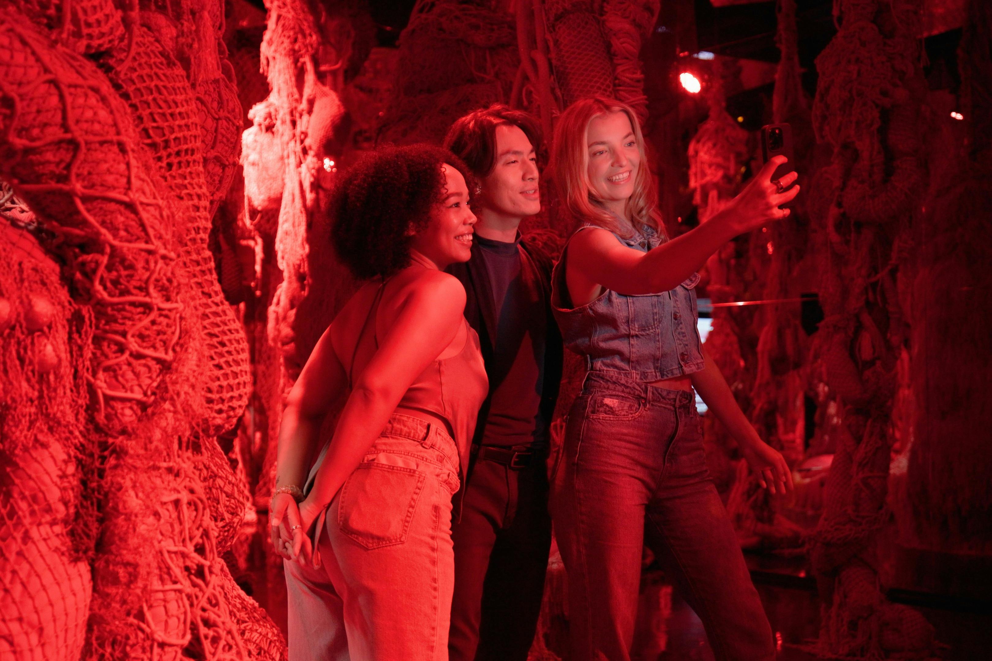 Three friends take a selfie in a dimly lit, red-hued room filled with hanging net decorations.