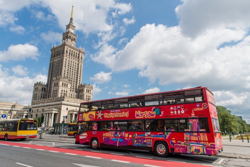 En röd dubbeldäckad City Sightseeing-buss i Warszawa med en hög historisk byggnad i bakgrunden under en delvis molnig himmel.