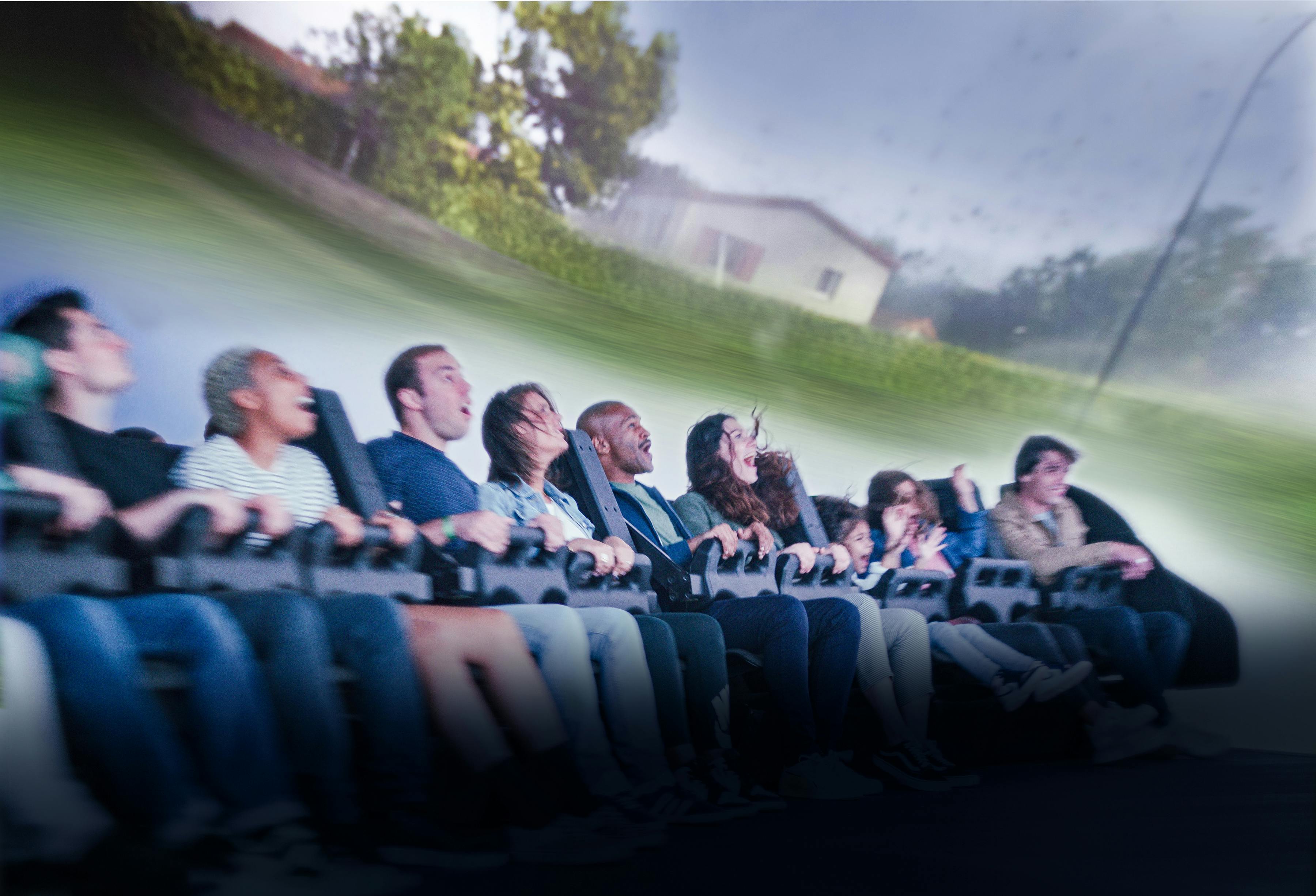 A group of people seated in a motion simulator ride experiencing a scene with projected visuals of a countryside.