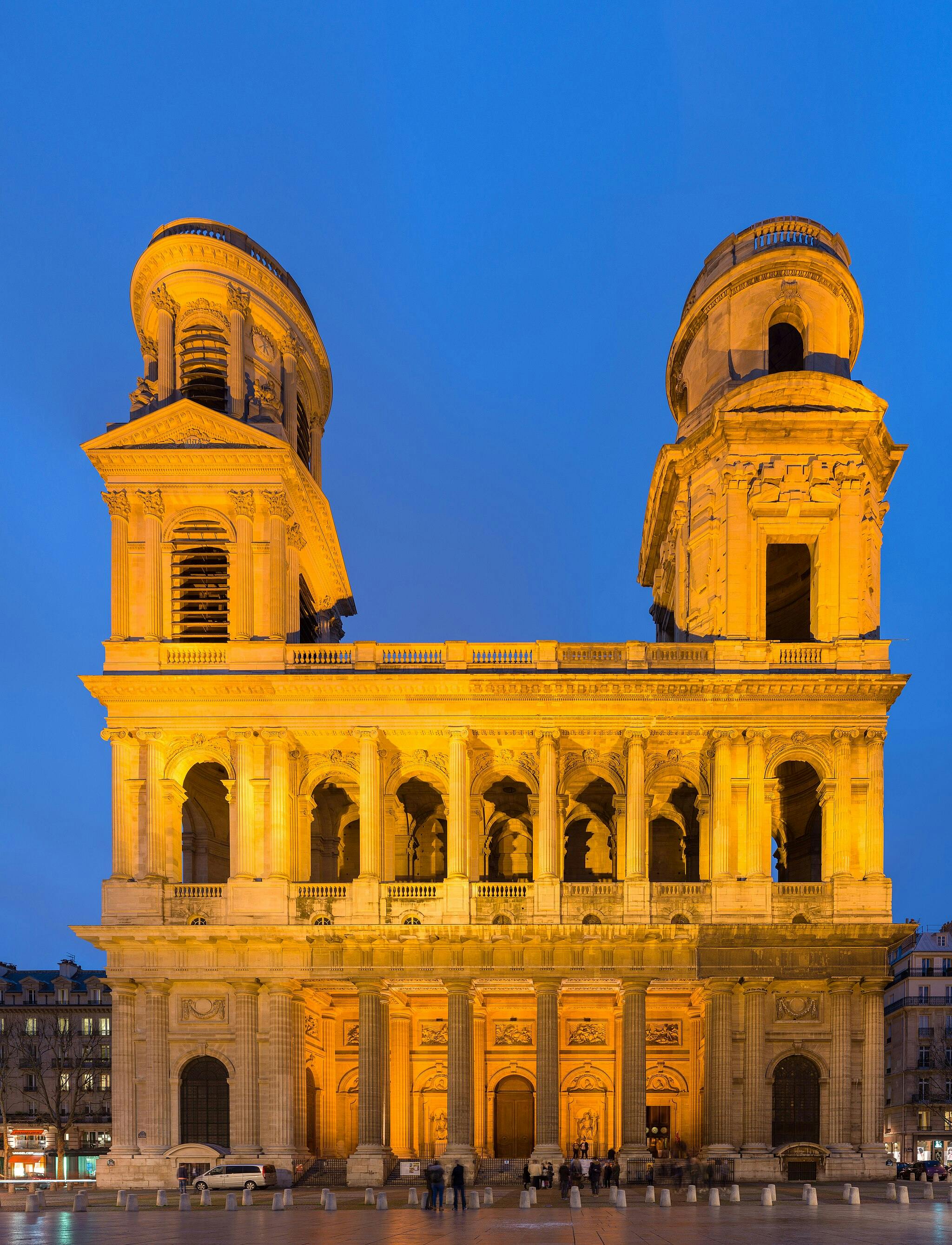 An illuminated, two-towered neoclassical building with arches and columns, against a twilight sky. People are gathered at the entrance.