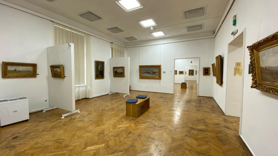 A spacious art gallery with paintings on white walls, parquet flooring, and a wooden bench with blue cushions in the center.