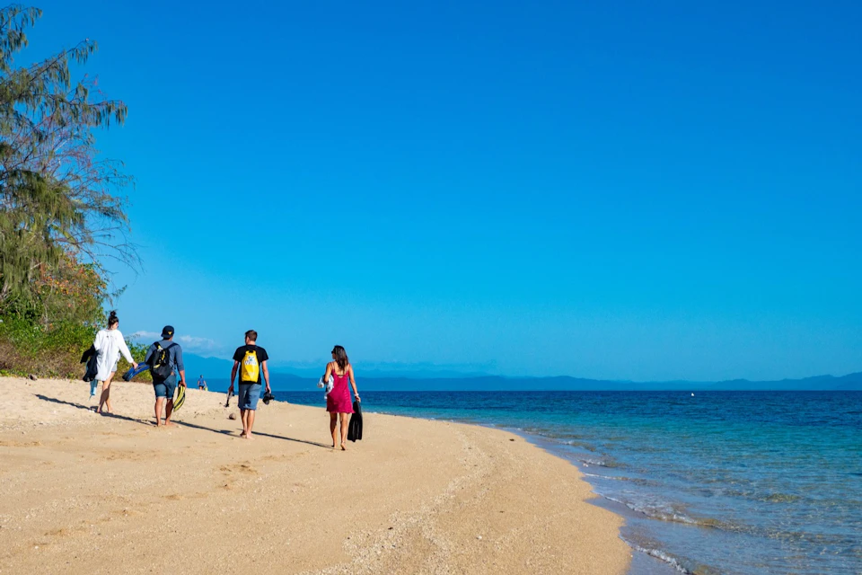 Four people walking on a sandy beach with calm, blue ocean water and clear sky in the background.