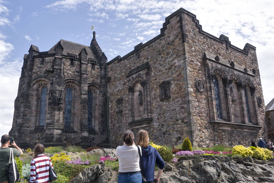 Two people stand facing a historic stone building with ornate architecture, surrounded by flowers and a rocky landscape.