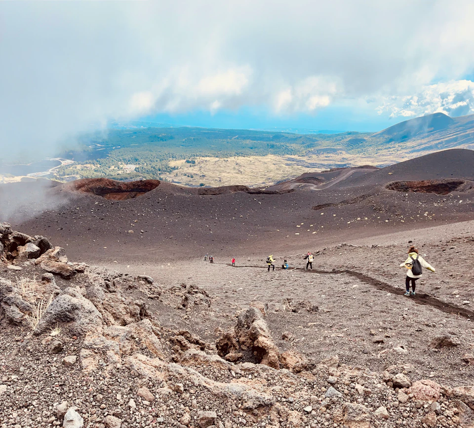 People hiking down a rocky, volcanic slope with a distant view of plains and mountains under a cloudy sky.