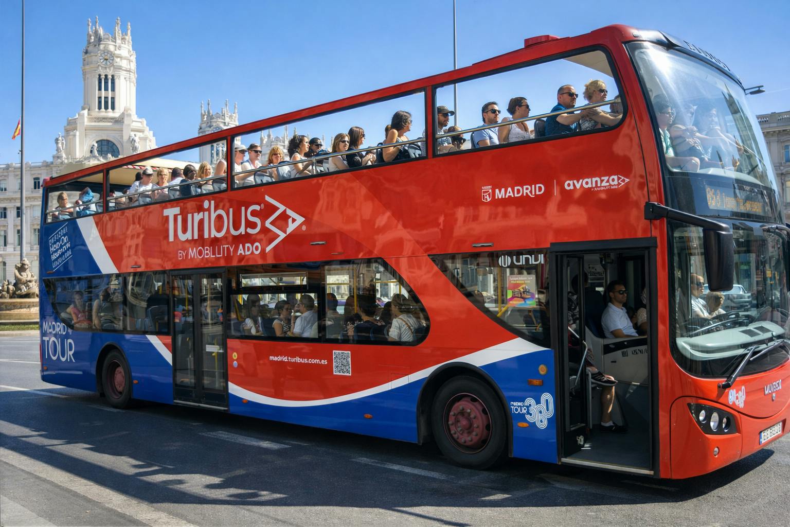 A red double-decker tour bus with passengers seated on both the upper and lower decks, parked at a stop in a sunny city.