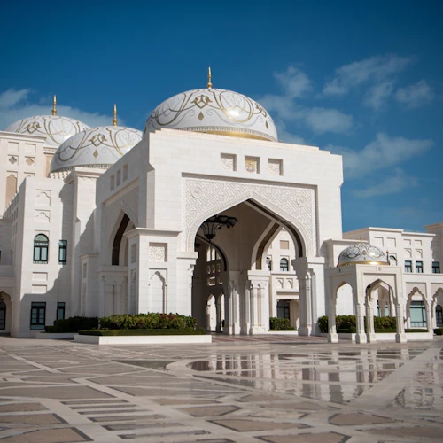 Verziertes weißes Gebäude mit mehreren Kuppeln, verschlungenen Bögen und einem geräumigen, gefliesten Innenhof unter strahlend blauem Himmel.