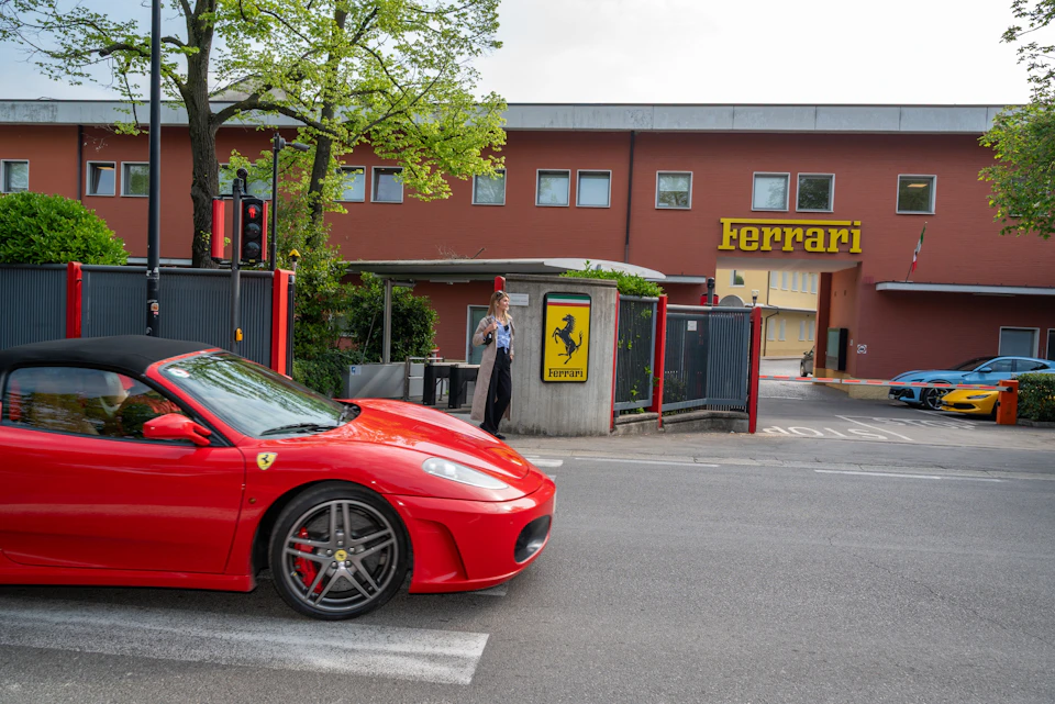 A red Ferrari car passing in front of a building with a Ferrari logo and a pedestrian nearby.