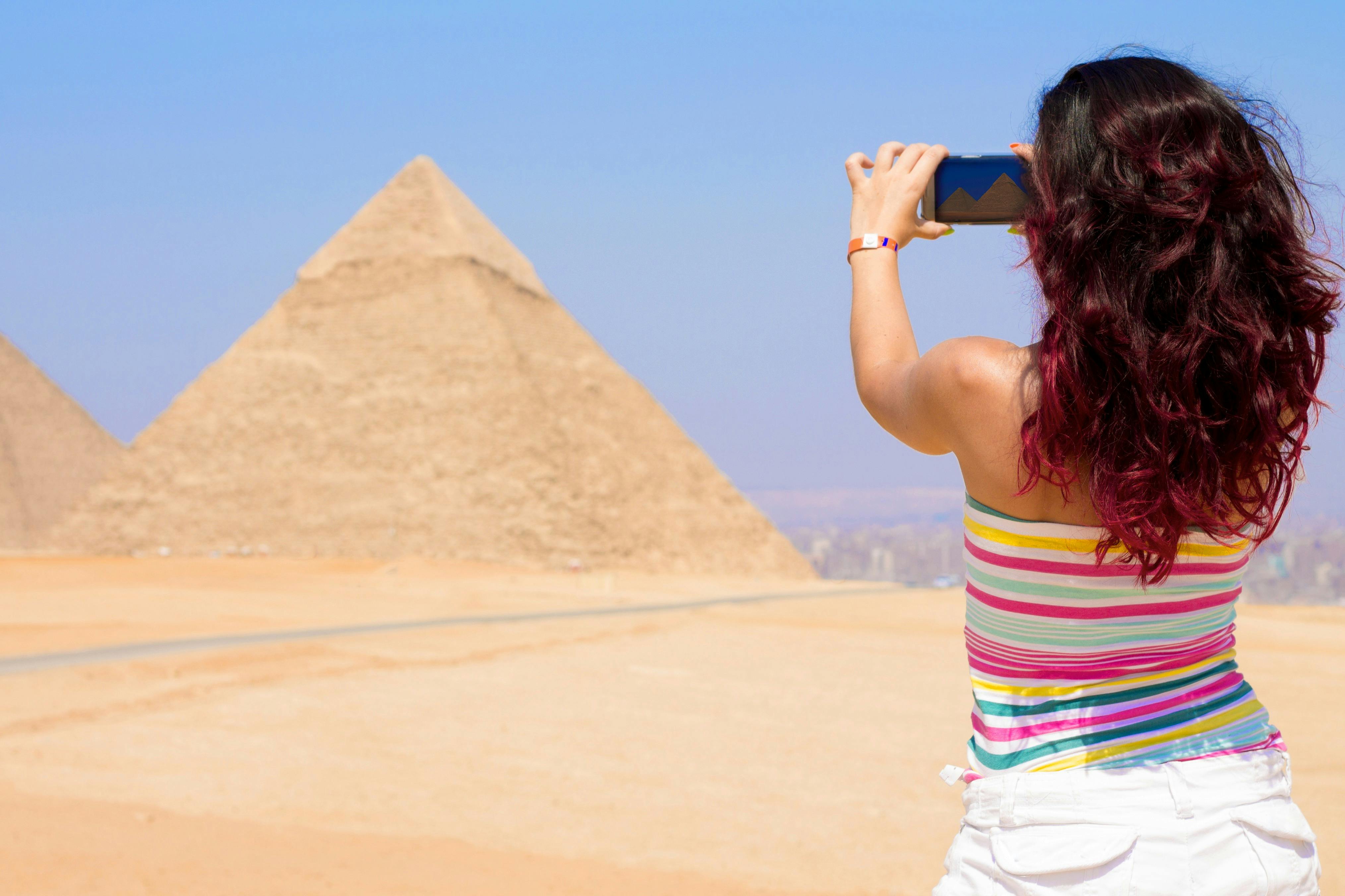 A woman takes a photo of the Great Pyramid with her phone. She wears a colorful striped top and stands in a sandy desert.