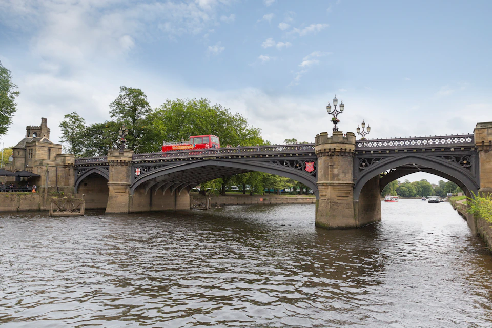 A red double-decker bus crosses an ornate bridge over a river with trees in the background and a partly cloudy sky.