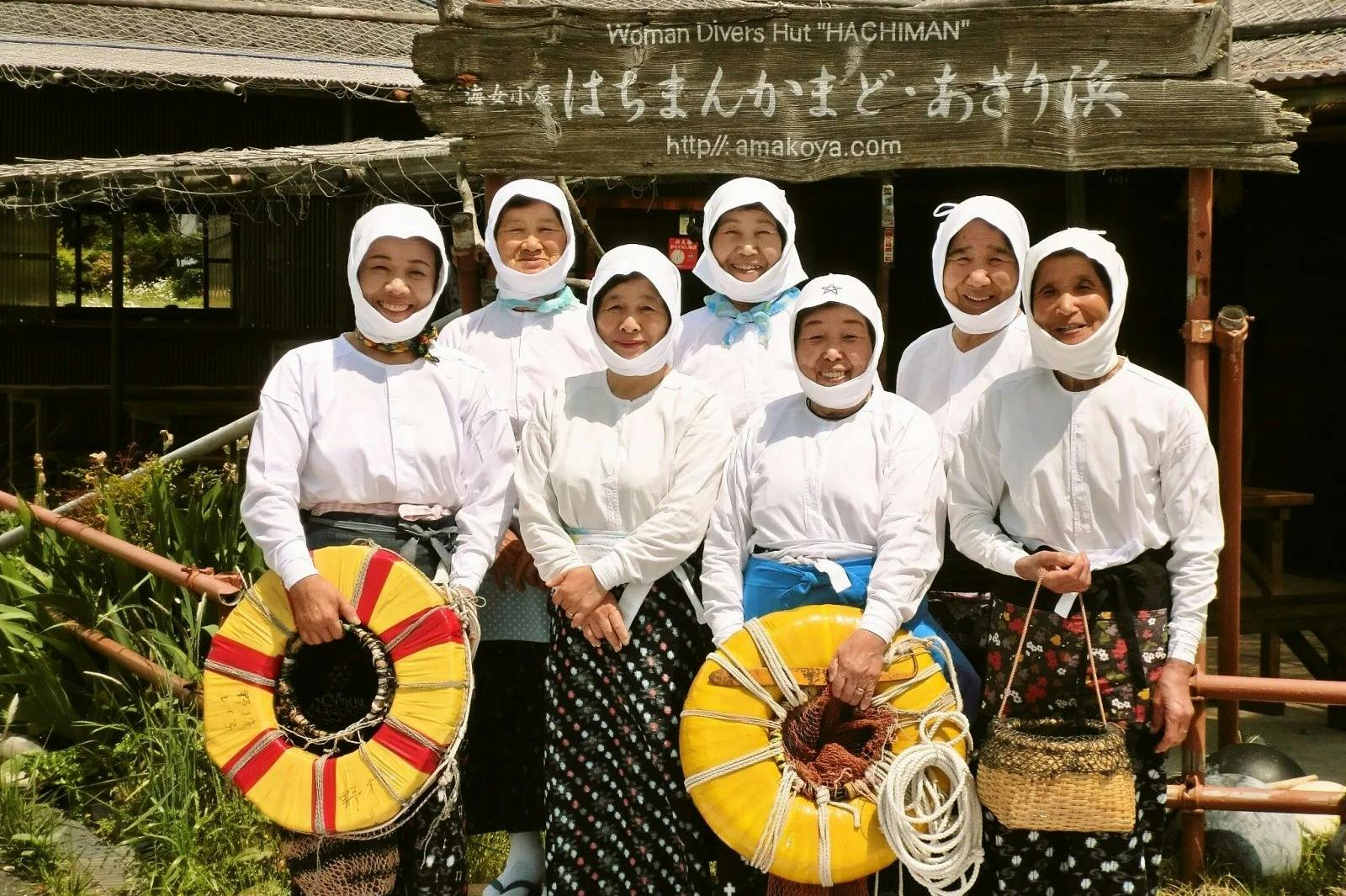 전통 복장을 한 8명의 여성 그룹이 "Woman Divers Hut 'Hachiman'" 의 나무 표지판 앞에서 다채로운 부유 장치를 들고 포즈를 취하고 있습니다.