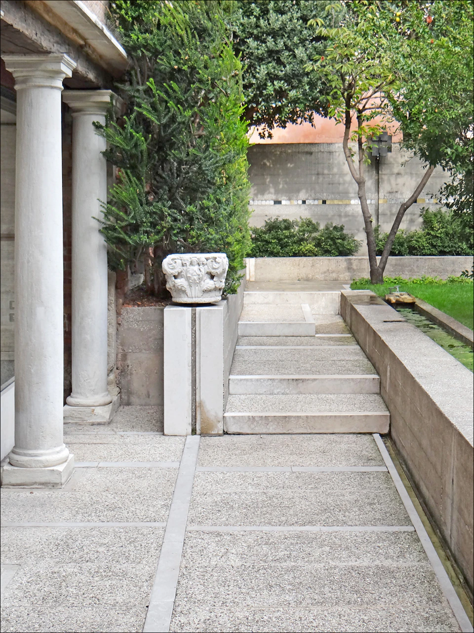 Stone-paved pathway with steps, bordered by greenery, a tree, columns, and a sculpted stone ornament, leading to a wall.