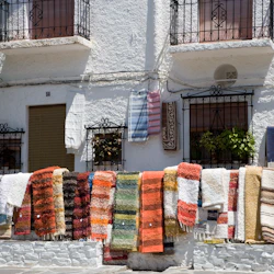 Färgglada mattor som hänger på en vit vägg med järnbalkonger och fönster i bakgrunden. En person är delvis synlig till vänster.