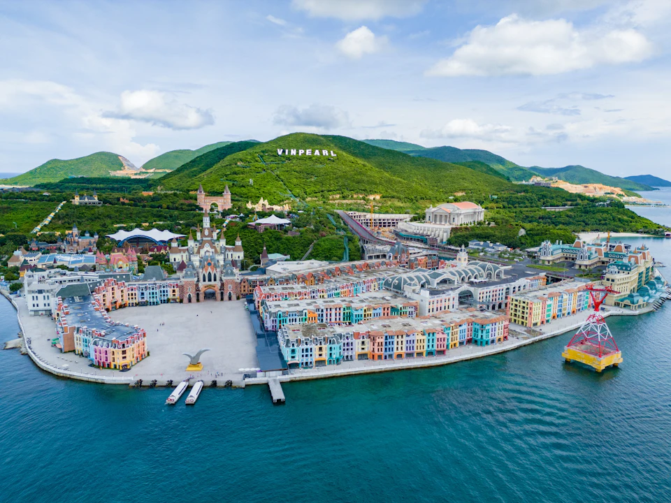A colorful town by the water with a green hilly backdrop and 'VINPEARL' sign on the hill under a partly cloudy sky.