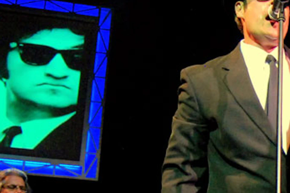 A band performing on stage with a singer in a suit and hat, accompanied by a guitarist. Images of two men in sunglasses are displayed behind them.