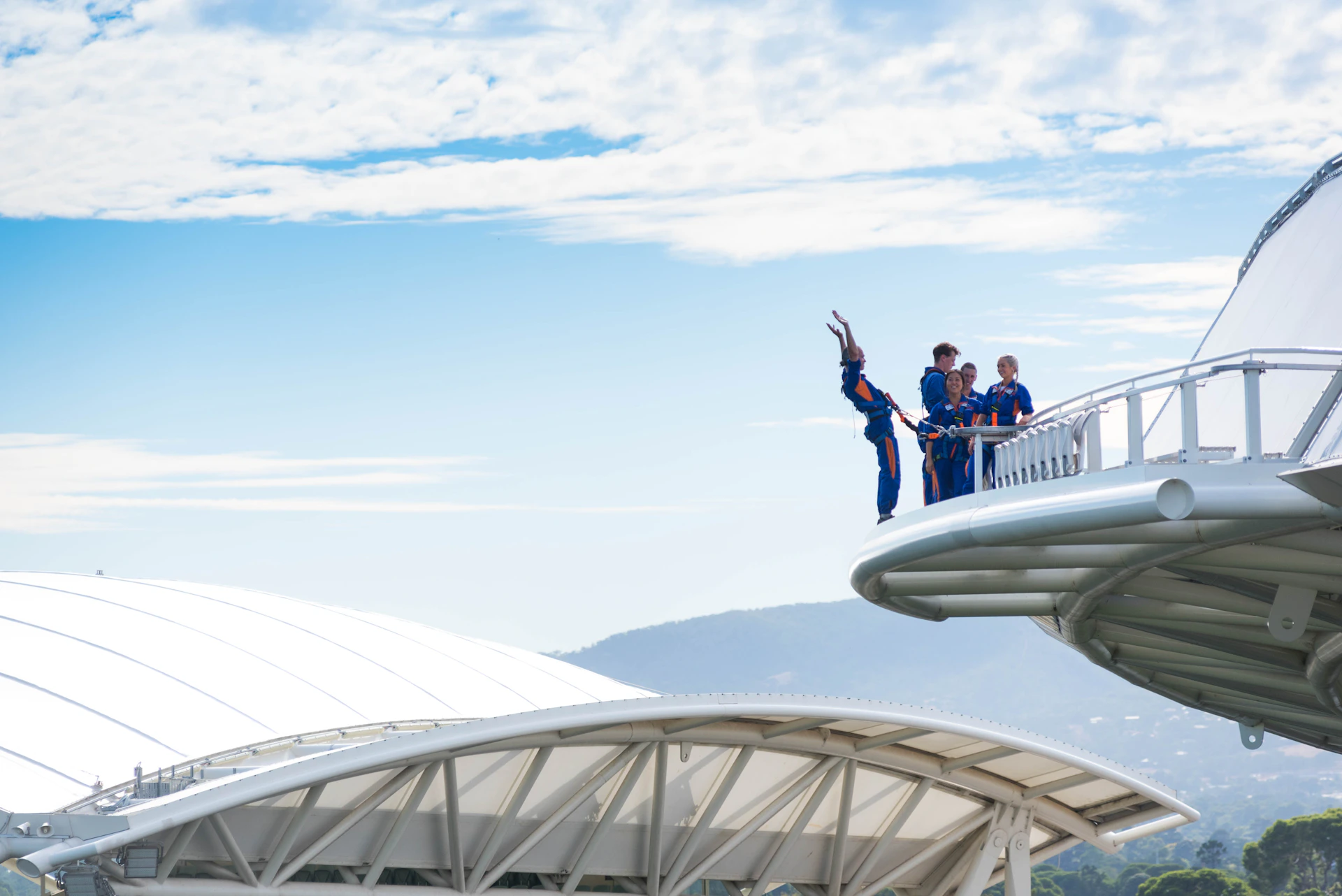 People in blue suits on a platform, one jumping off with hands up, with a background of a domed structure and a partly cloudy sky.