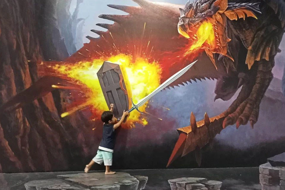 A child interacts with a 3D mural of a dragon breathing fire, holding a shield and sword as if defending against it.